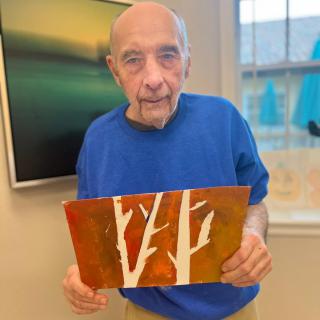 An elderly man in a blue shirt holding a colorful painting of trees indoors.