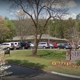 A parking lot with multiple cars in front of a building surrounded by trees, featuring a sign in the foreground.