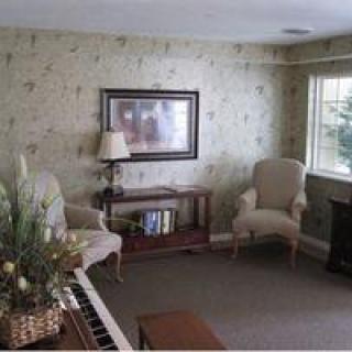 A cozy living room with a piano, armchairs, and a bookshelf near a large window.