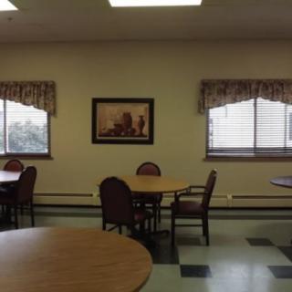 A quiet dining area with several tables and chairs, featuring windows with curtains and a framed picture on the wall.