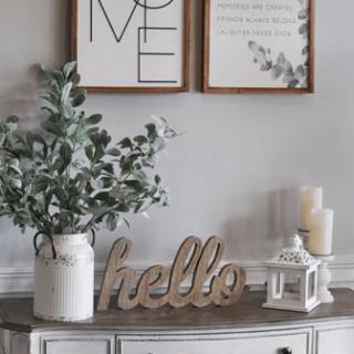 A cozy home decor setup featuring a vase with greenery, a welcoming 'hello' sign, and candles on a stylish console table.