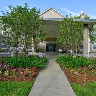 A landscaped garden leads to the entrance of a building, flanked by tall trees and vibrant flowers.