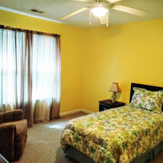 A cozy bedroom with yellow walls, featuring a single bed covered in a floral patterned duvet. The room includes a chair, a small bedside table with a lamp, and is illuminated by natural light through the window with sheer curtains.