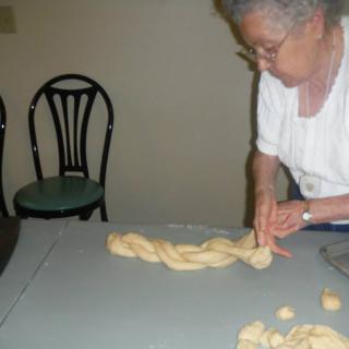 An elderly woman braiding dough on a kitchen table.