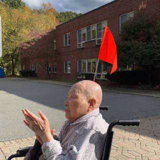 An elderly person in a wheelchair enjoying the outdoors, with a red flag visible against a backdrop of autumn foliage and a building.