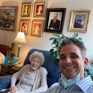 A man takes a selfie with an elderly woman seated in a living room filled with framed pictures on the wall.