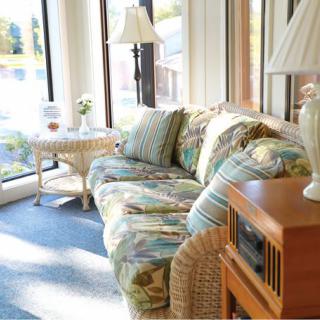 A sunlit room with a floral-patterned sofa and cushions next to large windows, featuring a lamp and a small table.