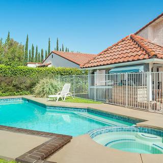 A tranquil backyard oasis featuring a swimming pool next to a house, enclosed by a safety fence with lush hedges in the background.