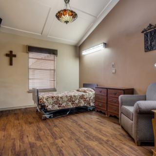 A hospital room with a single bed, wooden dresser, and armchair, featuring a cross on the wall and a window with blinds.
