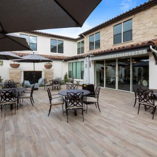 A spacious outdoor patio area with tables, chairs, and umbrellas in front of a modern building with a stone facade.