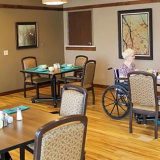 An elderly person in a wheelchair sits at a table in a dining room with multiple tables and chairs.