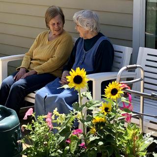 Two elderly women sit and chat on a bench in a garden surrounded by flowers.