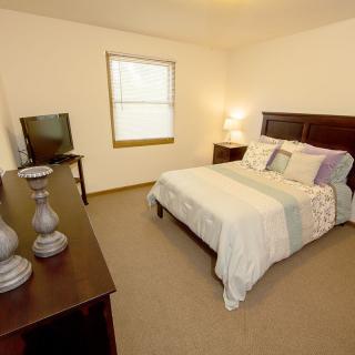 A cozy bedroom featuring a bed with decorative pillows, a dresser with a TV, and a warmly lit lamp.