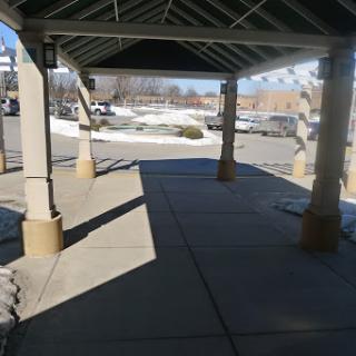 A covered walkway with concrete pavement and shadows, leading to a parking lot.