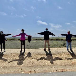 A group of people standing outdoors, posing with outstretched arms under a clear sky.