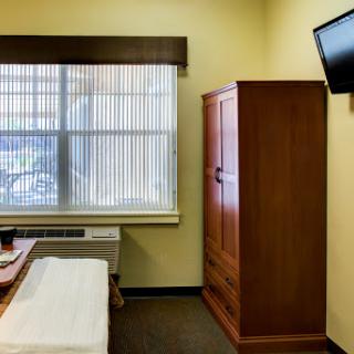 A neatly arranged hotel room featuring a wooden wardrobe, a television mounted on the wall, and large windows with curtains.