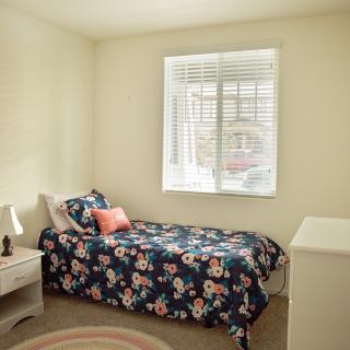 A cozy bedroom with a single bed featuring floral bedding, a small lamp on the nightstand, and a window letting in natural light.