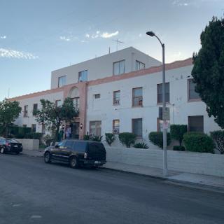 A view of a residential building along a street, with parked vehicles and green shrubbery.