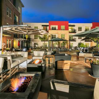 A modern outdoor patio area with fire pits and comfortable seating, set against an apartment building backdrop at dusk.