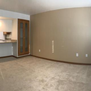 An empty room with beige walls and carpet flooring, featuring a view into a kitchen area.