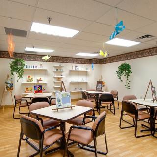 A cozy room with tables and chairs, decorated with plants and art supplies on shelves.