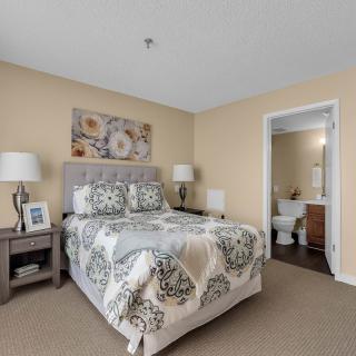 A cozy bedroom with a neatly made bed, decorative pillows, and bedside lamps. The room also features a carpeted floor and wall art, with a bathroom visible in the background.