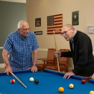 Two seniors enjoy a game of pool together in a room decorated with an American flag.