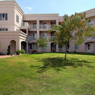 A sunny view of a three-story building with balconies and a well-maintained lawn with a tree in the foreground.