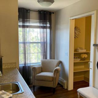 A cozy kitchen area with an armchair by the window, allowing sunlight to filter through sheer curtains. A small storage room with neatly arranged towels is visible beside the kitchen counter.