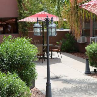 A tranquil patio setting with tables and chairs under umbrellas, surrounded by lush greenery and lit by a classic lamp post.