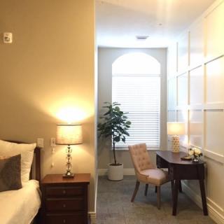 A cozy bedroom with a desk, chair, and decorative plant by a large window.