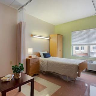 A clean and minimalist hospital room featuring a single bed, wardrobe, and window providing natural light.