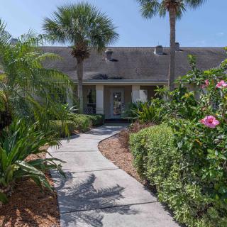 A charming house with a garden pathway surrounded by palm trees and blooming flowers on a sunny day.