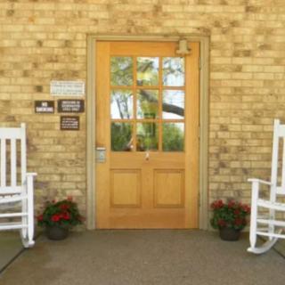 A wooden door with glass panels is flanked by two white rocking chairs and potted plants against a brick wall.