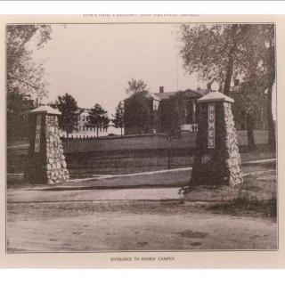 A historical black and white image of a gated entrance with stone pillars, flanked by trees and a fence.