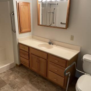 A bathroom with a wooden cabinet, sink, mirror, toilet, and shower featuring a grab bar.