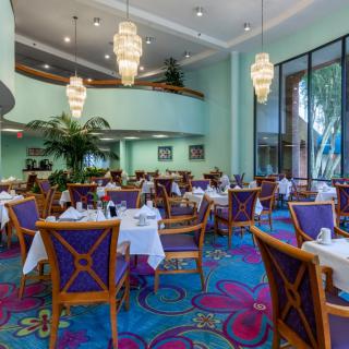 A spacious restaurant interior with elegantly set tables and colorful decor, featuring large chandeliers and a vibrant carpet.