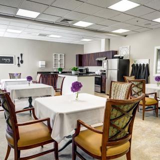 A modern dining area with neatly arranged tables and chairs, adorned with purple flowers, adjacent to a kitchen space.