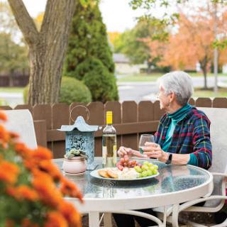 An elderly woman enjoys a serene moment outdoors, sipping wine and savoring snacks amidst the autumn scenery.