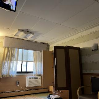 A view of a hospital room featuring a television, window with curtains, and an air conditioner.