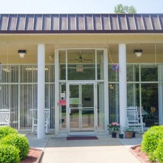 Front entrance of a building with glass doors, columns, and rocking chairs on a sunny day.
