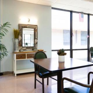 A modern living room with a wooden table, chairs, a decorative mirror, and large windows allowing natural light.