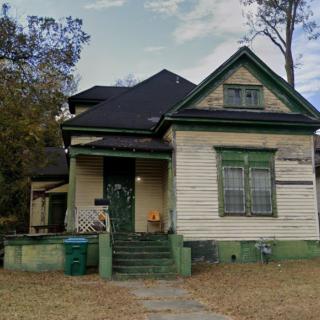 A historic wooden house with a small porch and triangular roof, surrounded by trees.