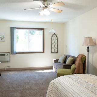 A cozy bedroom with a large window, armchair, and ceiling fan.