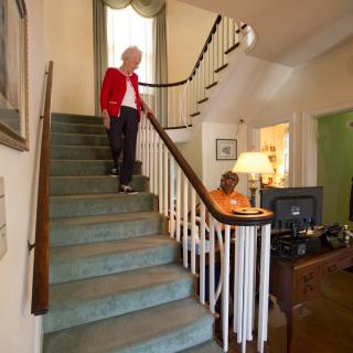 A person in a red jacket descends a staircase while another person sits at a desk in the living room.