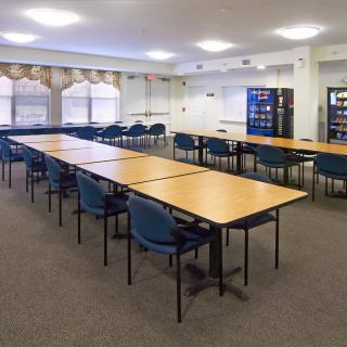 A well-lit conference room with tables arranged in a rectangular formation, surrounded by blue chairs and featuring vending machines along the wall.