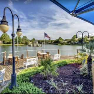A serene waterfront patio with a garden and seating area overlooking a tranquil lake under a clear blue sky.