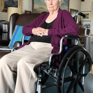 An elderly woman sitting in a wheelchair, looking thoughtfully ahead.