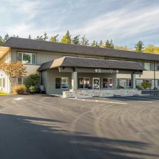A two-story motel with a parking lot in front, surrounded by trees under a clear sky.
