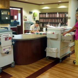 A nurse at a hospital reception desk with medical equipment in the foreground.